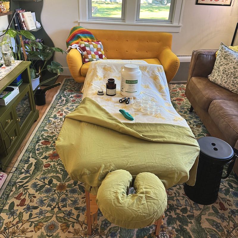 A massage table set up in a living room on a floral rug, between a mustard chair and a leather couch