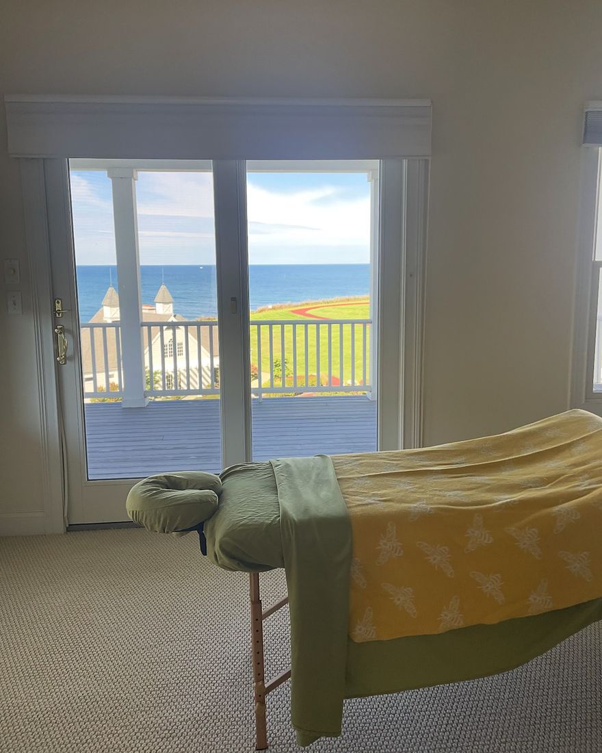 A massage table made up with sage and yellow linens in front of glass doors that look out over the water